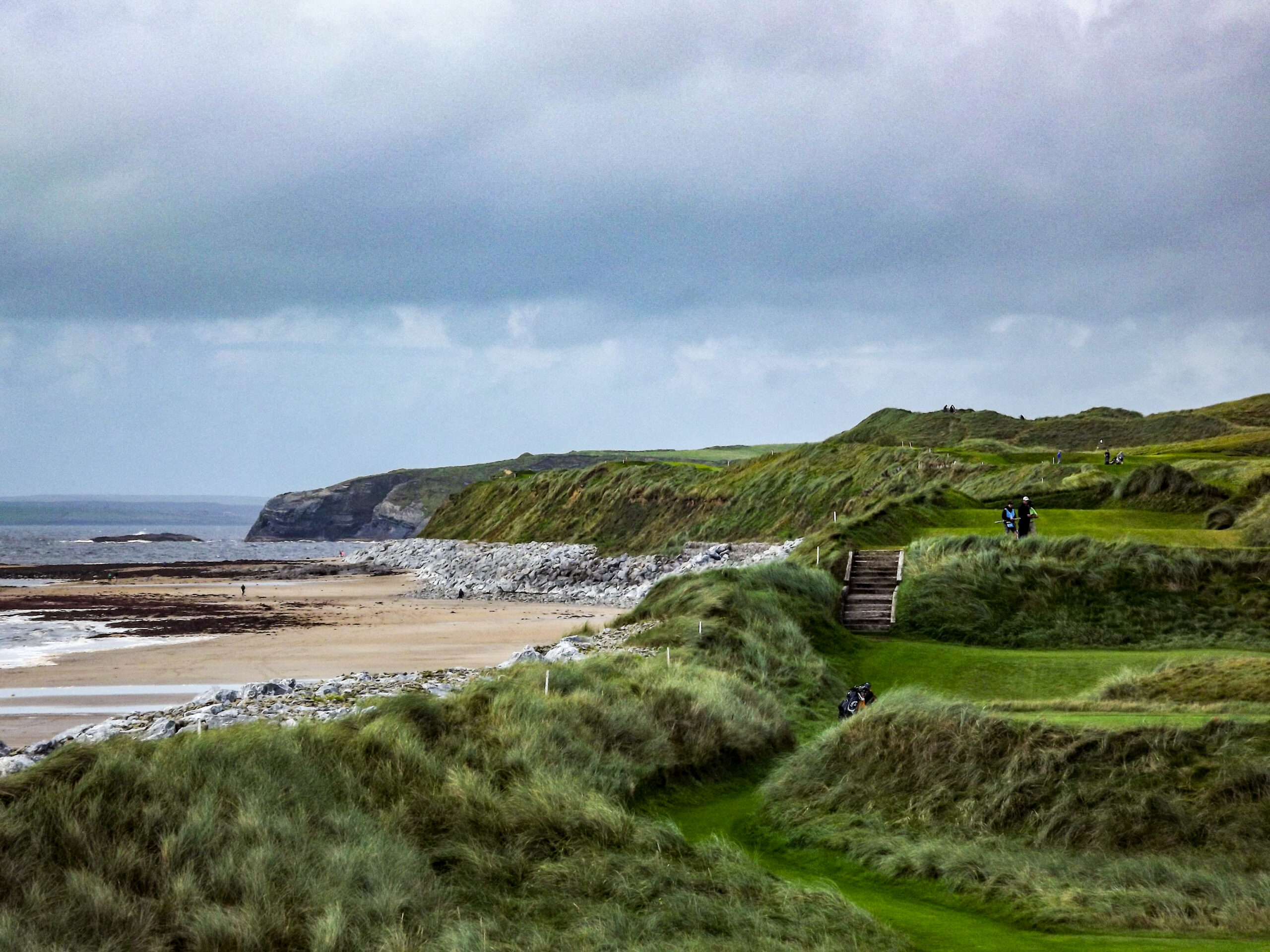 A scenic photo of the renowned Ballybunion Golf Course in County Kerry, Ireland, with the Atlantic Ocean waves crashing against the shore on the left, and the lush green fairways and rugged dunes stretching out to the right.