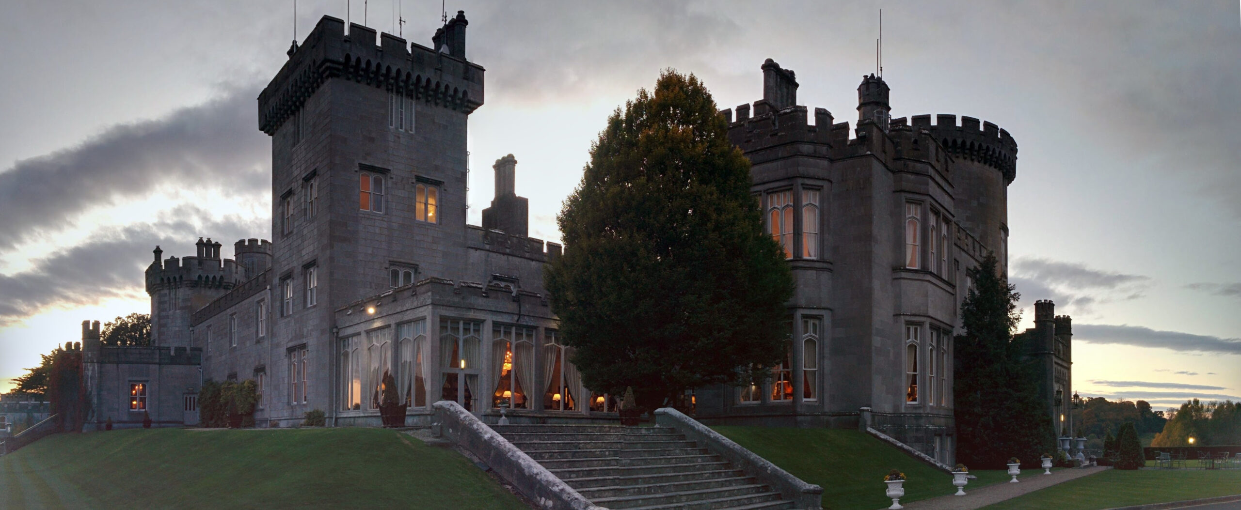 A breathtaking panoramic view of Dromoland Castle in County Clare, Ireland, at dusk, featuring the castle's majestic Gothic Revival architecture, beautifully illuminated against a warm, golden sunset, with lush green gardens. 