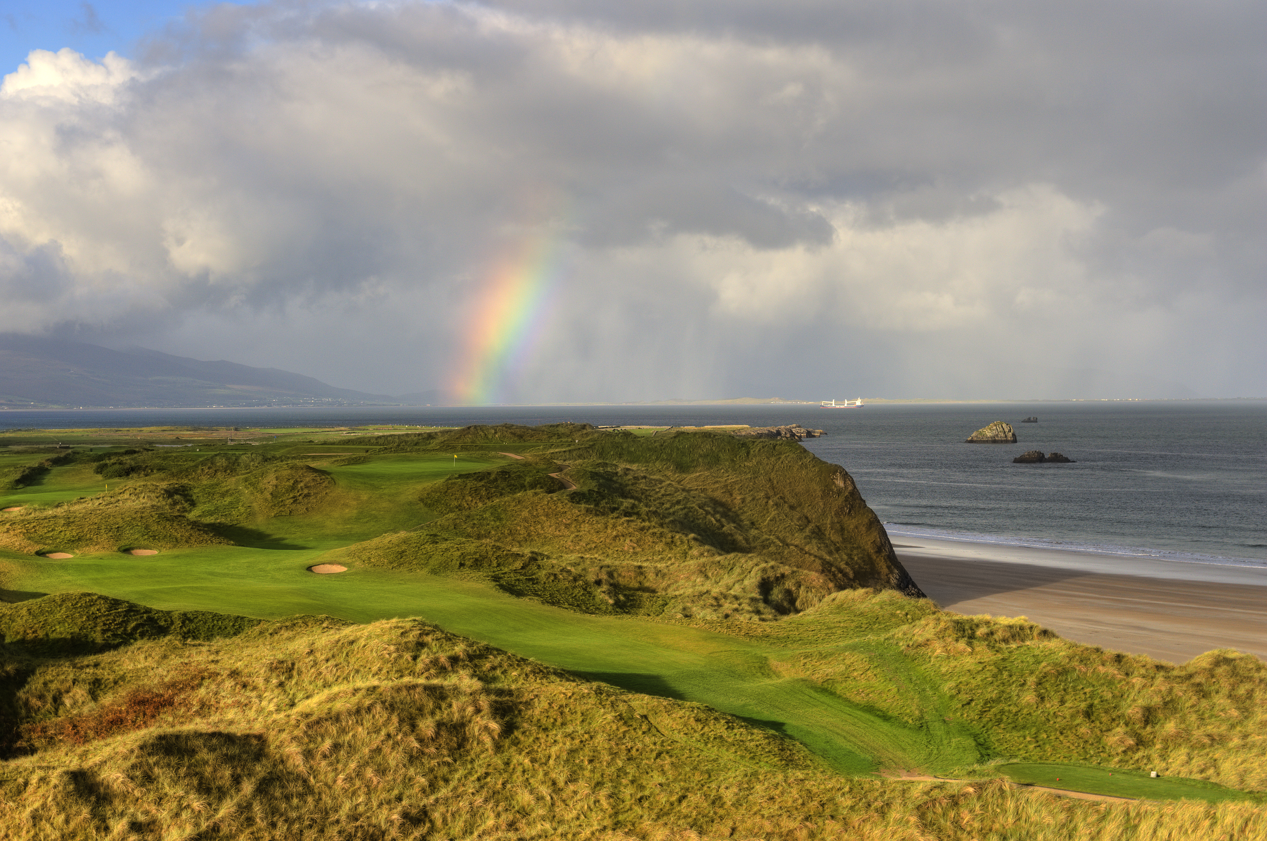 A scenic photo of the Tralee Golf Course in County Kerry, Ireland, showcasing its lush green fairways, rolling hills, and stunning ocean views along the rugged coastline.