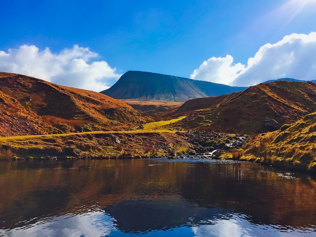 A breathtaking photo of the Brecon Beacons, a majestic mountain range in south Wales, UK, showcasing rolling hills, rugged peaks, and serene lakes, bathed in a warm, golden light, with a few wispy clouds scattered across the sky.