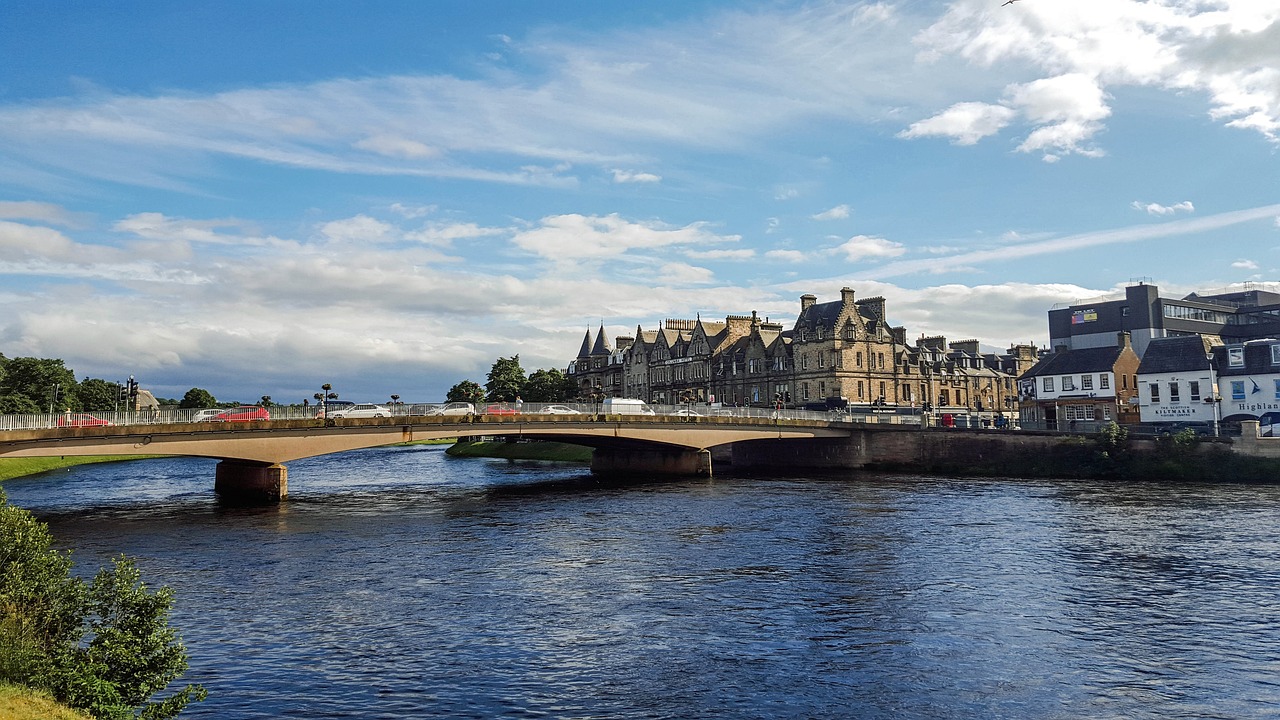 A picturesque stone bridge spans a serene river in Inverness, Scotland, surrounded by lush greenery and trees, with a few buildings and hills visible in the background. 