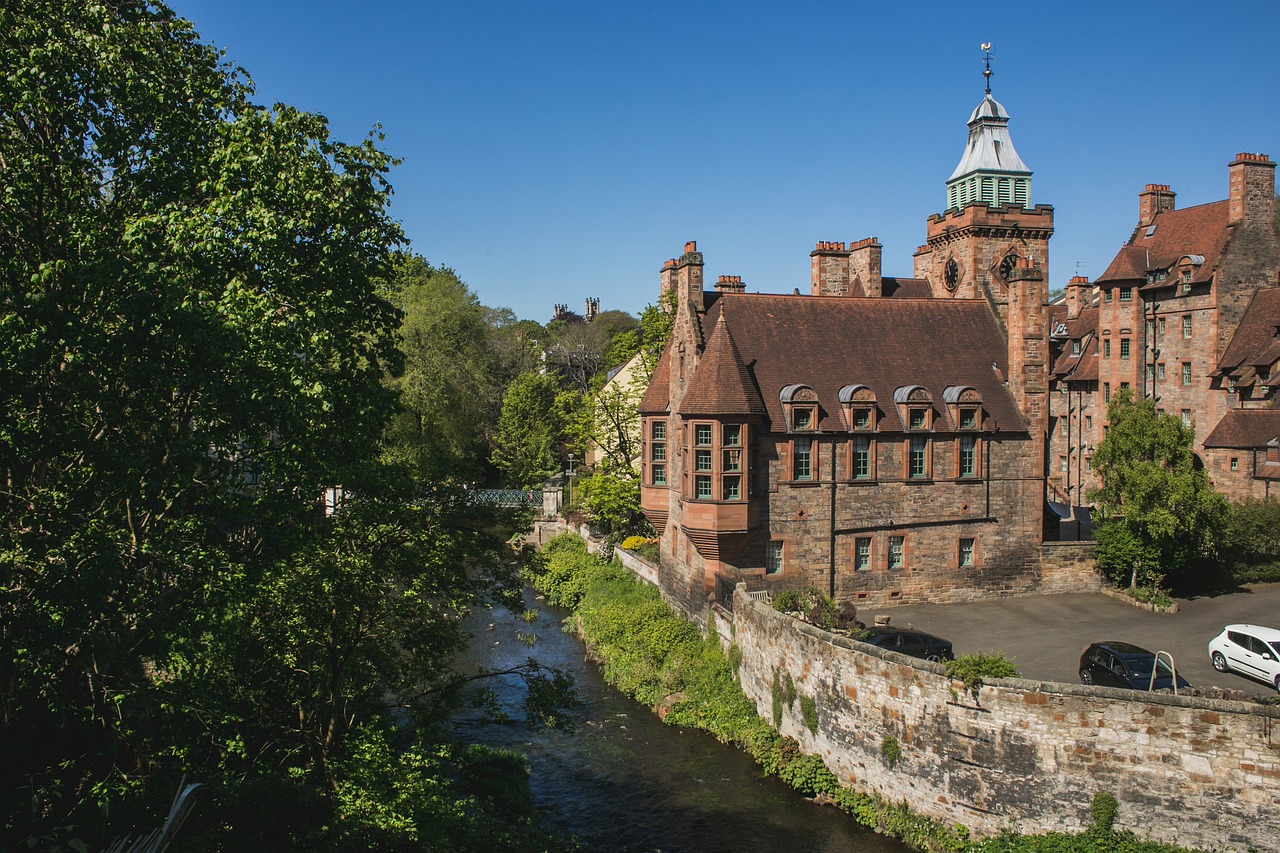 A charming and historic Dean Village in Scotland, featuring a picturesque stream running through the village, surrounded by quaint stone cottages, trees, and lush greenery. 