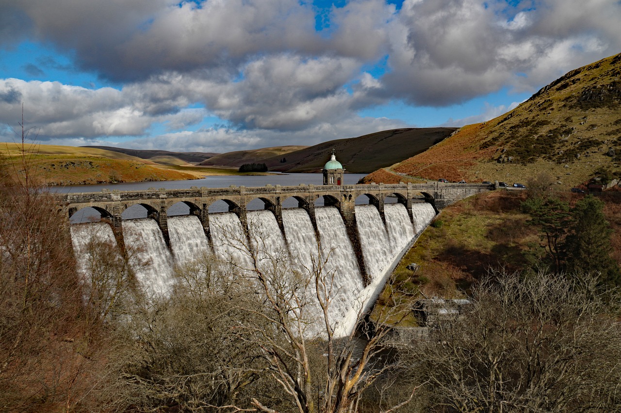 A serene photo of the Elan Valley Reservoirs, specifically Graig Coch, in mid-Wales, showcasing a tranquil lake surrounded by lush green hills, woodlands, and scenic walking trails, with the distant mountains rising majestically in the background.