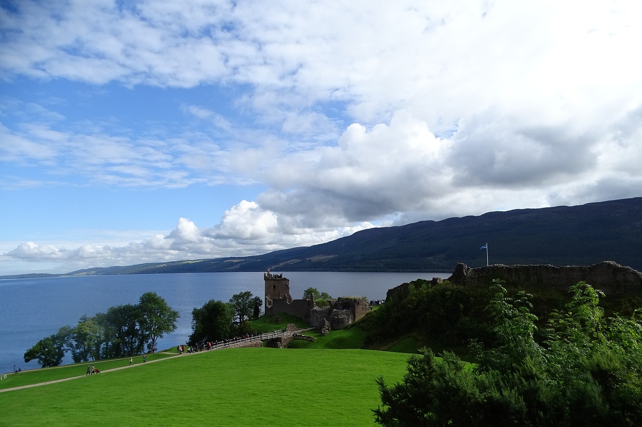 A serene and misty Loch Ness in Scotland, with calm waters reflecting the surrounding rolling hills and trees, under a soft and cloudy sky. 