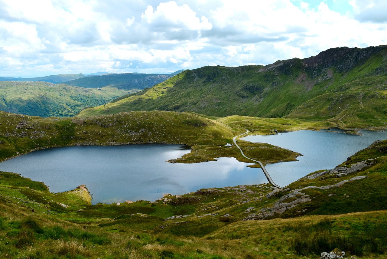 A breathtaking photo of Snowdonia National Park in Wales, UK, showcasing a stunning mountain landscape with rugged peaks, sparkling lakes, and lush green valleys, with a few wispy clouds scattered across the sky, capturing the park's natural beauty and majesty.