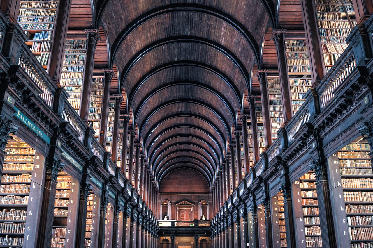 The historic Long Room of Trinity College Library in Dublin, Ireland, featuring a grand, high-ceilinged chamber with marble busts of famous scholars and rows of old, leather-bound books shelved in ornate wooden bookcases. 