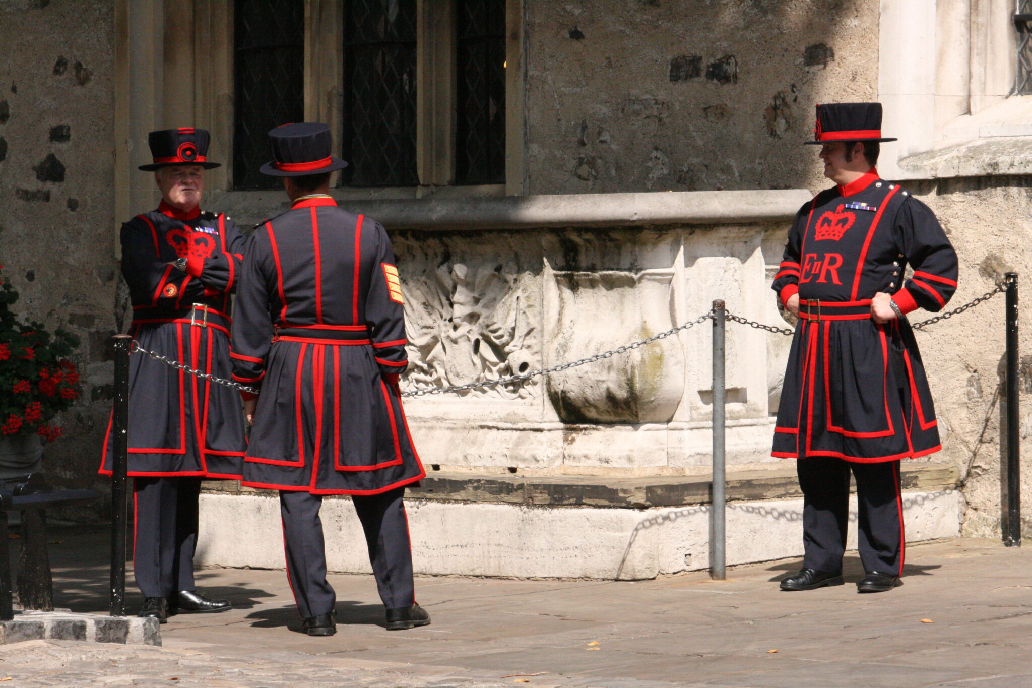 Three Yeoman Warders, commonly known as Beefeaters, stand in conversation, dressed in their traditional black and red ceremonial uniforms, adorned with gold braiding and medals, within the historic walls of the Tower of London.