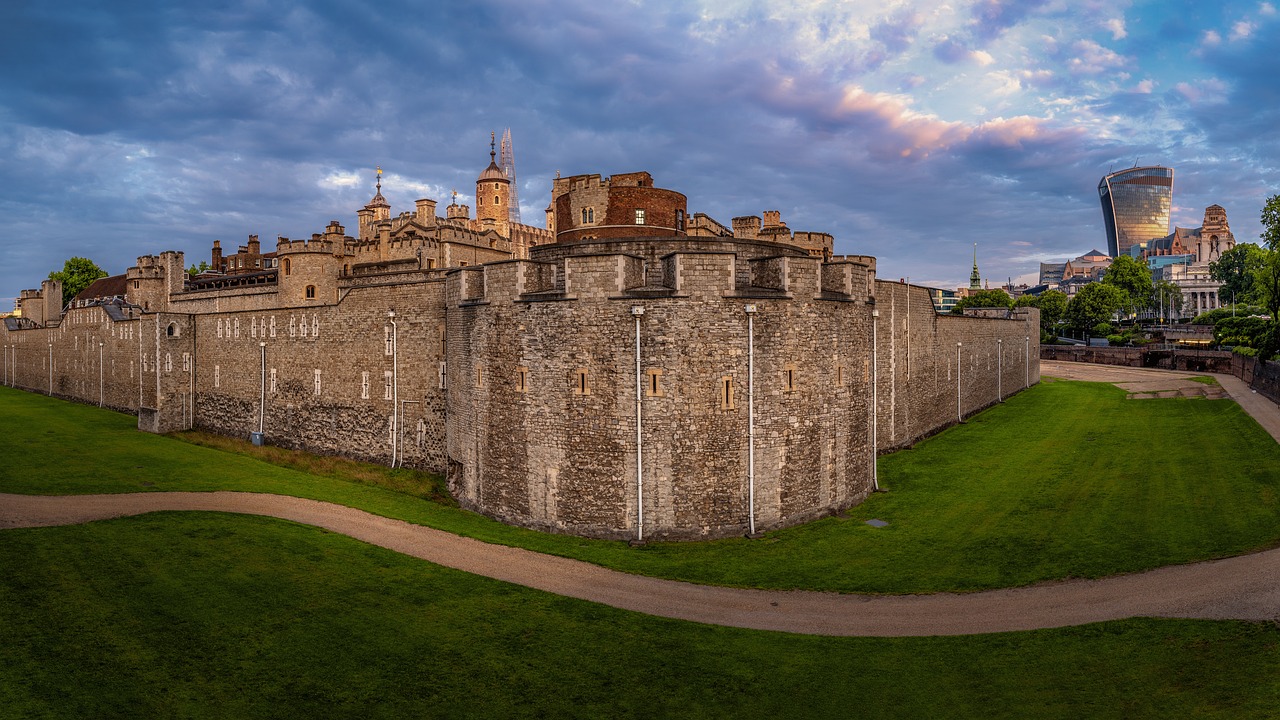 The Tower of London at sunset, with its medieval walls and towers bathed in warm golden light, surrounded by lush greenery and the bustling cityscape of London.