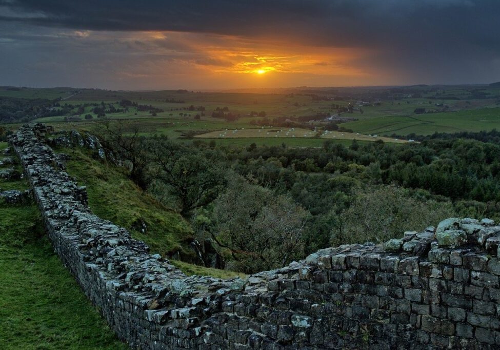 Hadrian's Wall at sunset with vibrant orange and pink hues illuminating the ancient Roman fortification, set against a serene countryside backdrop.